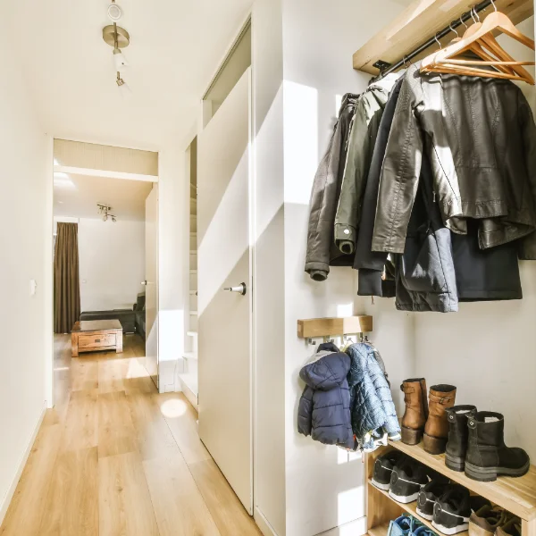 Bright entryway mudroom featuring jackets hanging on a wooden rack, boots neatly arranged on a lower shoe shelf, and a long hallway leading into the living room.