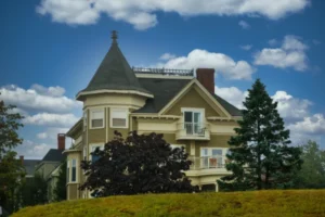 Large historic Victorian home featuring a corner turret, olive green siding, and balconies, situated on a grassy hill against a blue sky.