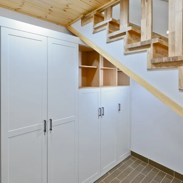 Custom built-in white storage cabinets and open wooden cubbies installed beneath a wooden staircase.