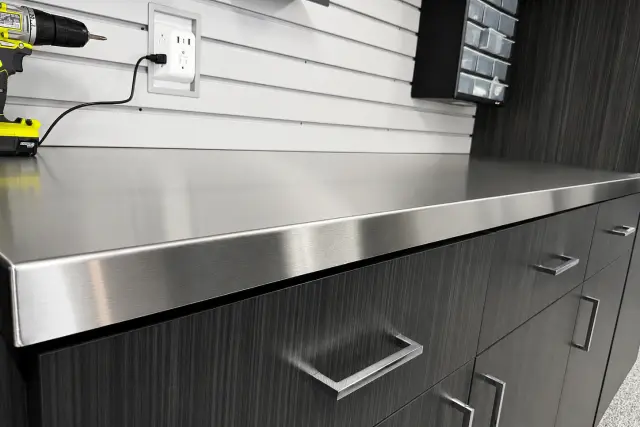 Close-up of a sleek stainless steel garage workbench countertop with integrated power outlet and slatwall storage.